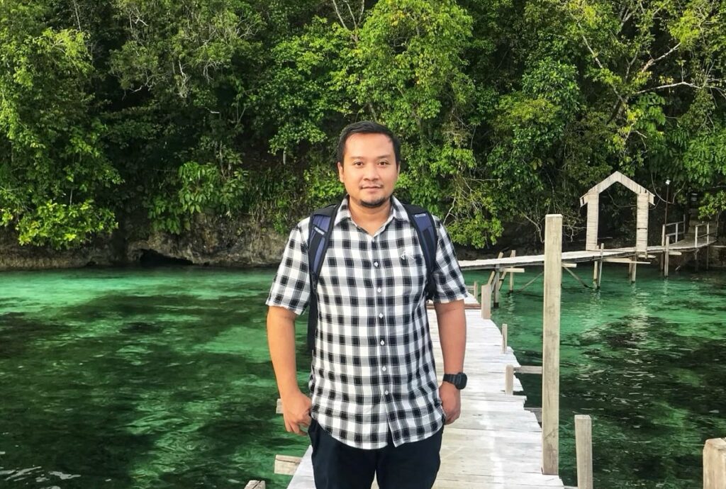 Whatsapp image 20260606 at 20 12 23 catrawarta Man in a black and white plaid shirt with a backpack stands on a wooden pier over clear green water with dense trees in the background